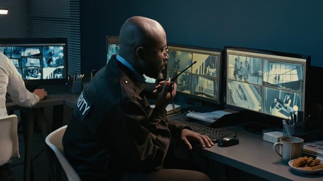 African American security officer watching TV footage and giving commands speaking into portable radio at night in control room, side view shot