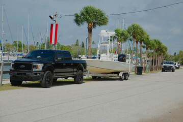 A black pickup truck parked at a marina towing a white center-console boat on a trailer along a palm-lined waterfront road under a cloudy sky.  © Del Harper