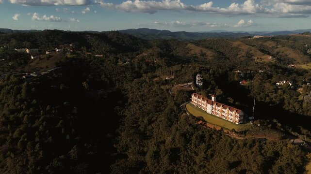 Aerial view of large European-style hotel building on hillside surrounded by forest and mountains with Capivari Park visible in background in Campos do Jordao, Brazil. Orbit camera movement