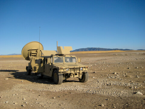 Desert-camouflage SatCom Humvee equipped with satellite communications dish parked in a remote desert landscape under a clear blue sky with mountains in the background. 