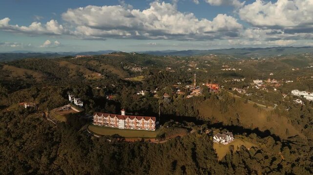 Aerial view of hotel building on hillside with camera moving toward hilltop where Capivari Park amusement park is located surrounded by forest and mountains in Campos do Jordao, Brazil. Push in camera