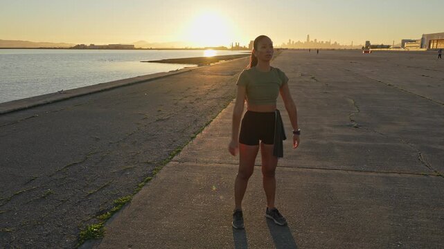 Fit young woman warming up and preparing for her running workout along a concrete path by the water, with the San Francisco skyline visible in the background during a beautiful golden hour