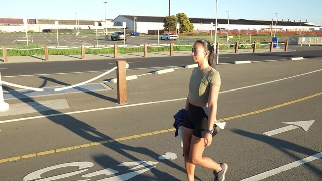 Determined young woman in sportswear walking through an empty parking lot after her workout, carrying a jacket and a water bottle while enjoying the warm light of the golden hour in Alameda