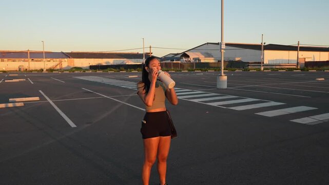 Thirsty young female runner in sportswear standing in a large empty parking lot during a golden sunset, hydrating herself with water from a reusable bottle after a long and hard workout