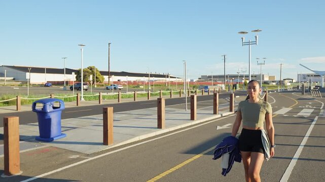 Young athletic woman in sportswear walking along a paved waterfront path in Alameda, California, after her morning run, with a determined expression and clear blue sky overhead