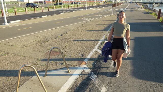 Active young woman in sportswear walking on a paved trail by the water after her morning workout, carrying a water bottle and a jacket while enjoying the sunrise in Alameda, California