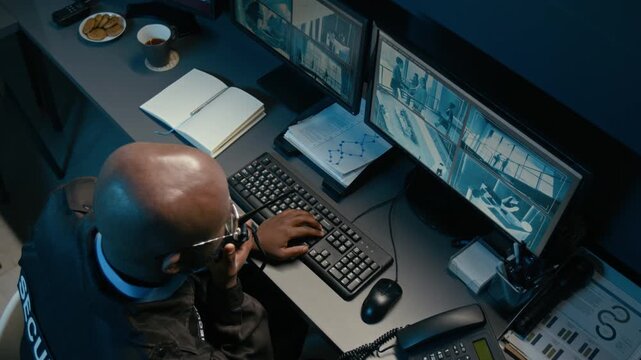 Top view shot of Black man watching computer monitors with CCTV footage while using portable radio and coordinating security operations from control room workstation during night shift