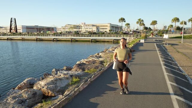 Athletic woman jogging towards the camera on a paved waterfront path during a sunny day. The active female runner is focused on her cardio exercise, enjoying a healthy outdoor lifestyle