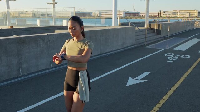 Young athlete walks along a waterfront path in an urban park, checking her fitness tracker while cooling down after an intense morning workout in the beautiful sunlight