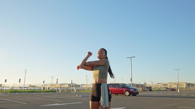 Young athletic woman warming up and stretching her body in an urban parking lot before her morning run, getting ready for a fitness workout while enjoying the bright morning sunshine