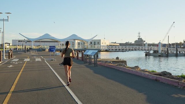 Beautiful woman with a long ponytail enjoying a morning run along the Alameda shoreline, with the waterfront and USS Hornet museum visible in the background against the bright sunlight
