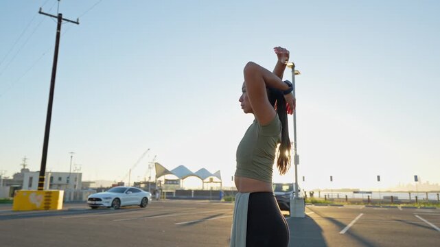 Fit young woman in sportswear stretching her arms and warming up before a run in an empty urban parking lot during a sunny day, getting ready for her workout and promoting an active lifestyle