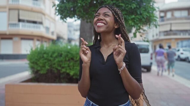 African american woman with fingers crossed and hands visible on a city street near buildings smiling with eyes closed; hope prayer luck.