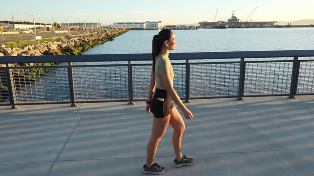 Side view of a determined fit woman in sportswear walking along a pier in Alameda, California, with the USS Hornet Museum visible in the background, enjoying a beautiful sunny day