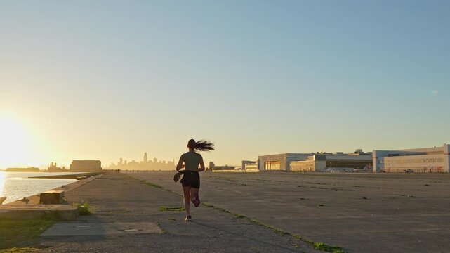 Young athletic woman running away from the camera on an old runway by the water at sunrise, with the San Francisco skyline visible in the distance, representing a healthy and active lifestyle