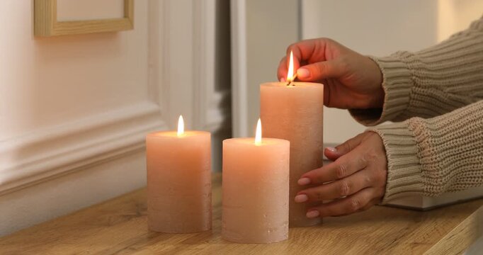 Woman lightning up candle at wooden chest of drawers with books near white wall indoors, closeup