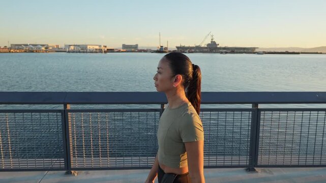 Young asian athlete trail running along the waterfront promenade in Alameda, California, with a view of the harbor and the USS Hornet Museum during a beautiful golden hour sunset