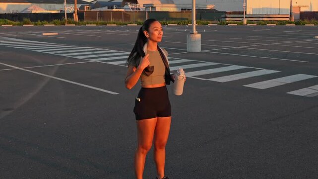 Tired female athlete drinking water from a bottle after a hard workout in an empty parking lot, getting ready to resume her jogging session during a beautiful and warm sunset in Alameda