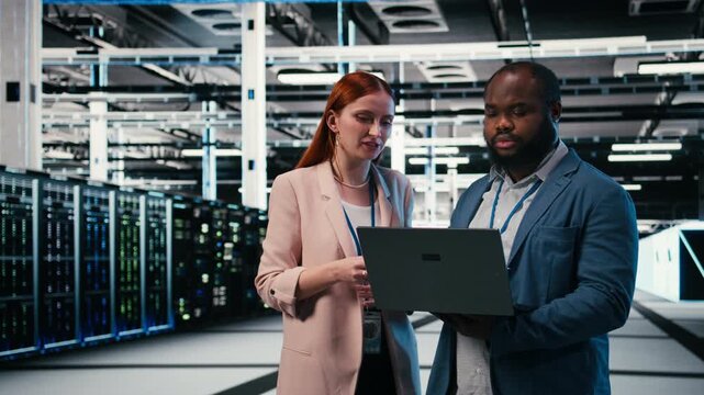 Optimizing electronics, laptop and manager teaching intern in server farm during induction. Monitoring system performance, notebook and team leader with trainee in data center, camera A