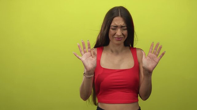 Young woman in studio with palms out and hands raised against lime green backdrop, squinting repulsed face wearing red crop top; disgust hesitation avoidance.