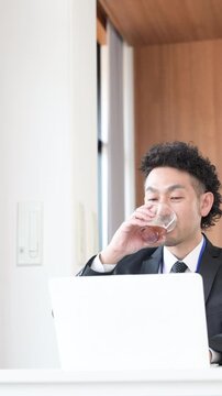 ノートパソコンを使い笑顔のビジネスマンの髭の男性　デスクで仕事をする縦動画　休憩でコーヒーを飲む