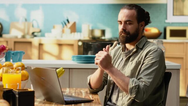 Adult male cracking his knuckles due to a nervous habit at home, making repetitive involuntary popping sound as he is focused on tasks. Using laptop at working in the kitchen, teleworking.