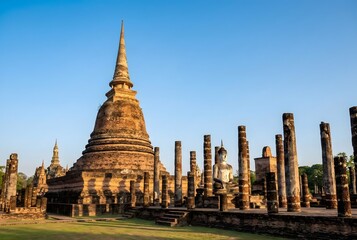 Majestic ancient Thai pagoda (Chedi) at Sukhothai Historical Park, ruins of stone pillars, serene atmosphere during golden hour, warm sunlight hitting the weathered brick texture, clear blue sky.
