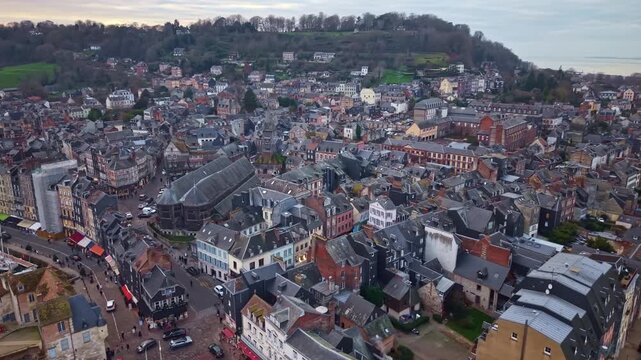 Dense cluster of dark slate-roofed timber-framed half-timbered buildings with white plaster walls, revealing distinctive wooden &Eacute;glise Sainte-Catherine church in Honfleur town France, drone orbiting