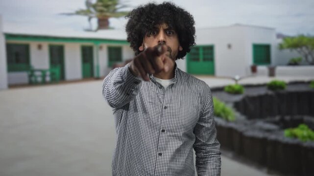 Man with curly hair and beard points finger at camera in white building courtyard outdoors, standing with tense posture; accusation.