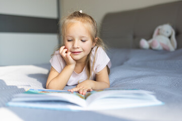 Fototapeta premium Young girl reading a book while lying on bed in a bright bedroom