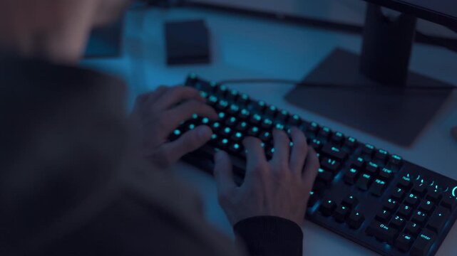 Hands typing on computer keyboard with blue backlight in modern workspace