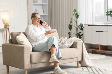 Obraz premium Smiling mature woman talking on smartphone while sitting on sofa in bright modern living room at home