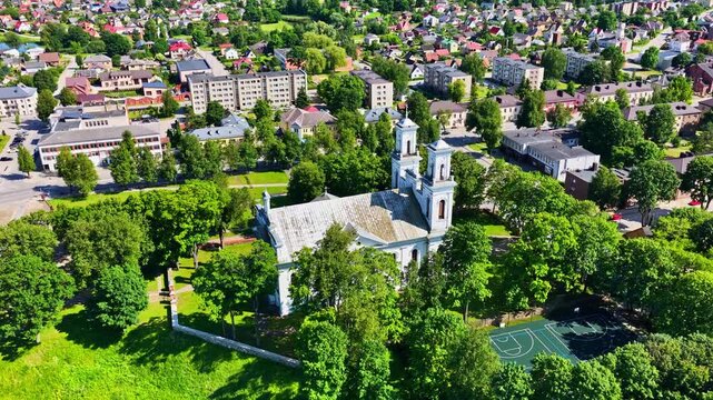 Drone aerial shot of Birzai St John the Baptist Church surrounded by lush green trees and nearby city buildings.