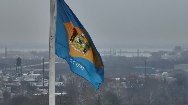 Close aerial drone view of Delaware state flag waving on a pole during cold winter weather with snowy landscape and distant city skyline visible through foggy atmosphere. Patriotic state in USA.