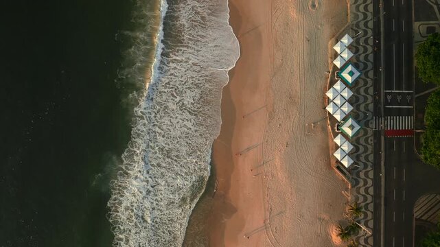 Top down aerial ascent over Copacabana beach waves, patterned sidewalk, and street during a sunny sunrise. Recorded in 4K Apple ProRes 422.