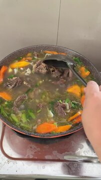 Indonesian beef cartilage soup or sup daging tulang rawan with carrots and potatoes simmering in rich broth. Woman hand stirring traditional soup in cooking pot on kitchen stove. vertical, closeup