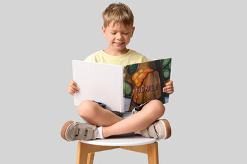 Fototapeta premium Cute little boy sitting on chair and reading book on grey background