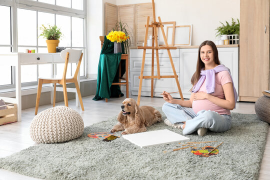 Young pregnant artist painting on floor with dog in room