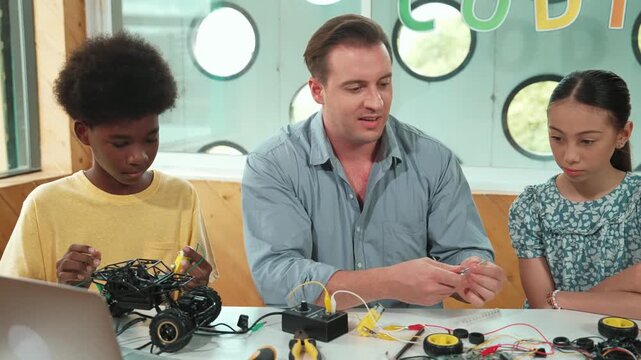Smart mentor teaching about electronic equipment while fixing car model. Caucasian teacher pointing while explain robotic construction at table with laptop, car model scatter around. Edification