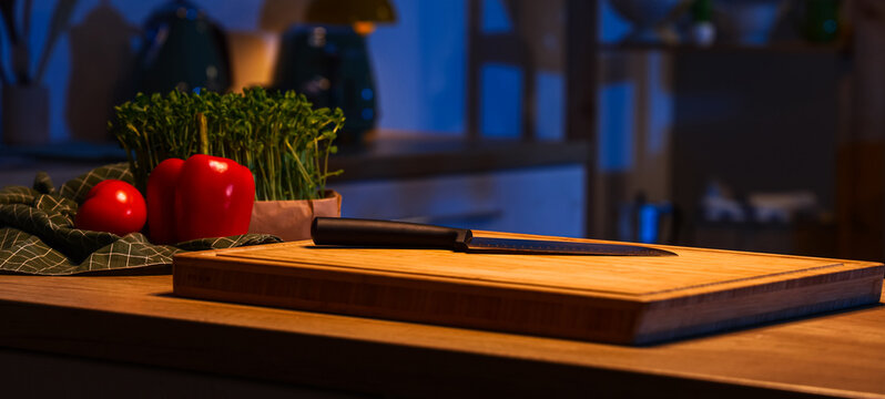 Cutting board with knife and vegetables on table in dark kitchen at night