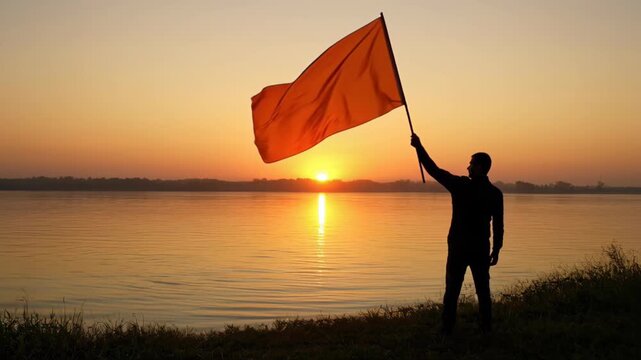 Silhouette of a Person Holding a Large Saffron Flag During Sunset by the Water's Edge