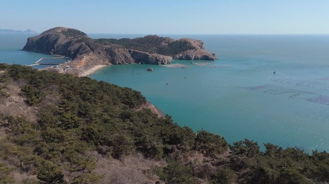 Aerial dolly-in video of a rugged peninsula on the coast near Dalian in northeastern China. The footage reveals steep rocky cliffs, a winding coastal road crossing the hill, and turquoise waters.
