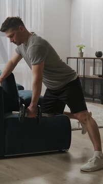 Focused home workout with a man performing dumbbell rows using a sofa for support in a bright living room, emphasizing strength, balance, and functional fitness training