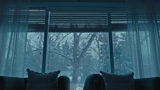 Static interior shot of living room with armchairs facing large window while snowstorm and snowy trees are visible outside.