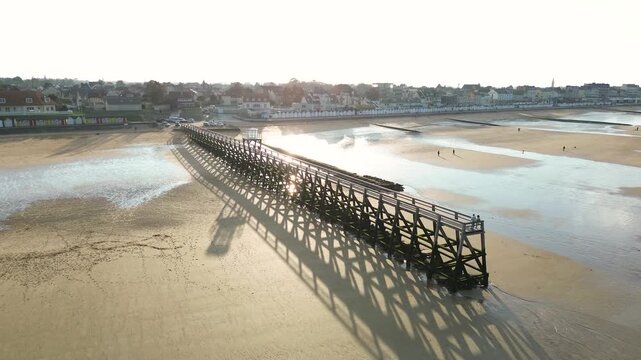 High aerial drone view of the coastline at Luc-sur-Mer in Normandy, France, during low tide. The wooden pier extends across wide tidal flats covered with seaweed and shallow pools reflecting the brigh