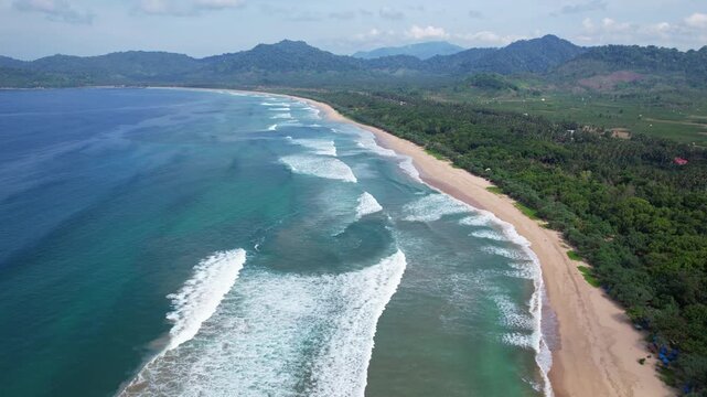 Aerial drone footage of the white sand Red Island beach bay, in Banyuwangi regency, Java island, Indonesia, with a beautiful crystal sea, many long waves, and a wide green forest behind