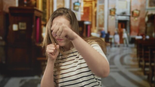 Woman holding fists raised near face inside a historic church with marble floors and wooden pews; determination.