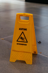 A yellow plastic warning sign on the floor with a German slippery caution message. The image represents safety, risk prevention, and public awareness.