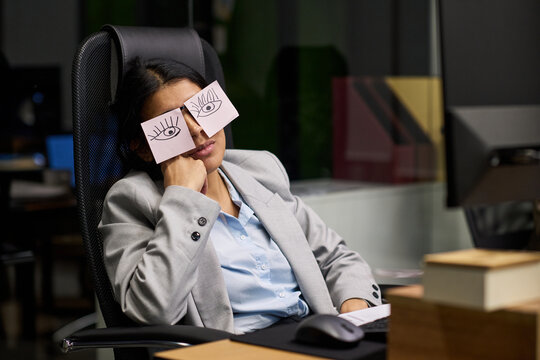 Hispanic woman sitting in office chair pretending to sleep at desk with paper drawings of eyes covering closed eyelids, demonstrating workplace fatigue
