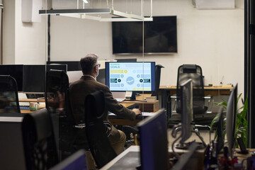 Fototapeta premium Middle aged Caucasian man sitting at desk working on computer with business analytics displayed on monitor in modern office environment, empty workstations in background